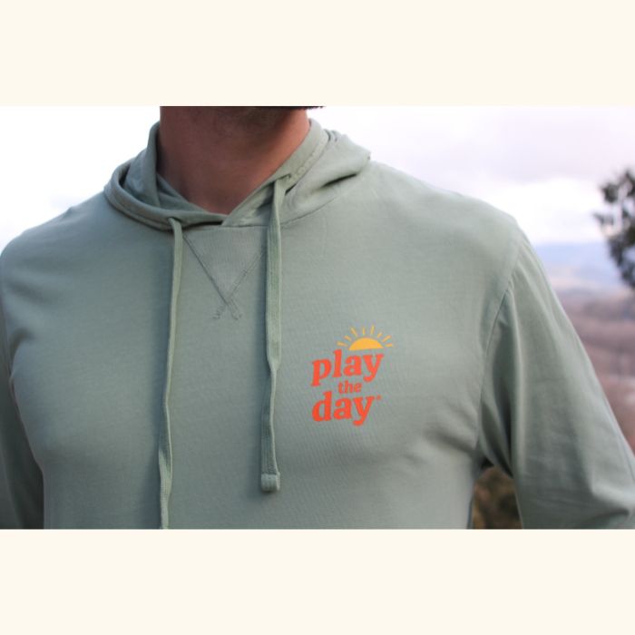 Close-up of Play the Day® logo on green long sleeve hooded tee made from soft cotton-blend fabric