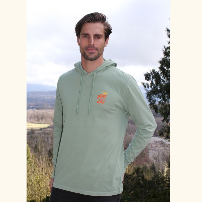 Man wearing green Play the Day® unisex long sleeve hooded tee with front logo, standing outdoors