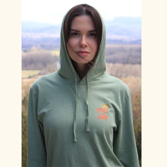 Woman wearing a unisex long-sleeved hooded tee in sage green with orange Play the Day® logo on chest