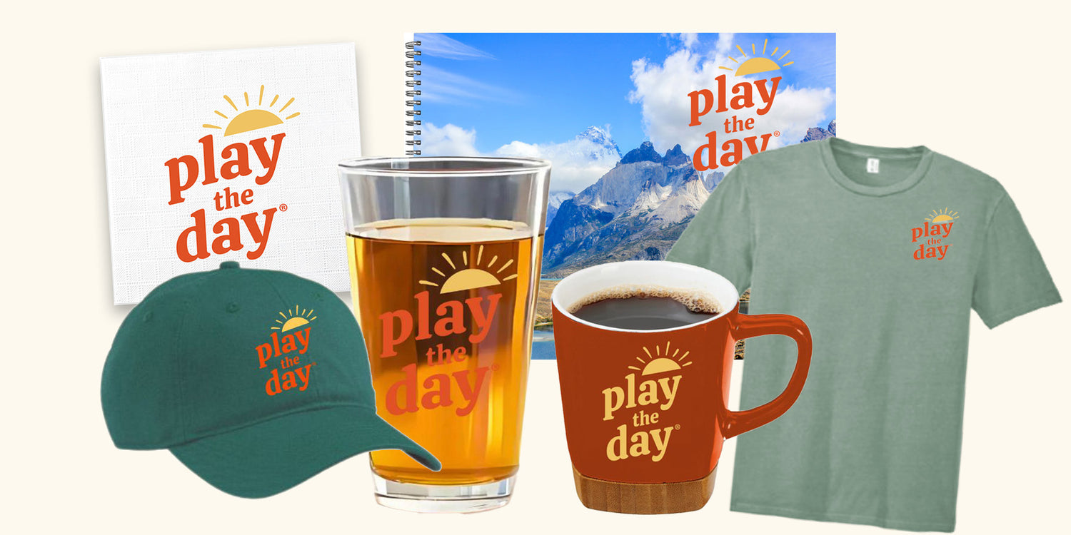 Smaller version of Play the Day® product collection including a journal, pint glass, t-shirt, hat, mug, and napkin, all featuring the Play the Day logo.