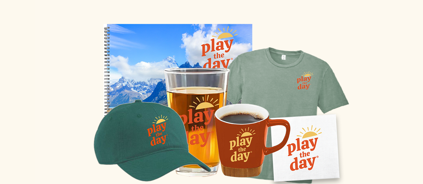 Play the Day® product collection including a journal, pint glass, t-shirt, hat, mug, and napkin, all featuring the Play the Day logo.