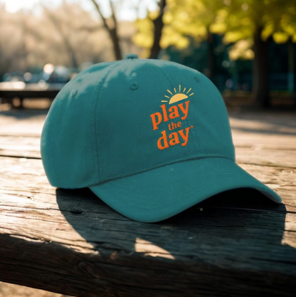 Play the Day® green baseball hat with orange logo resting on a wooden picnic table in warm afternoon sunlight.