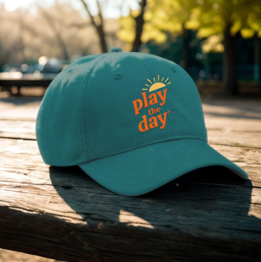 Play the Day® green baseball hat with orange logo resting on a wooden picnic table in warm afternoon sunlight.