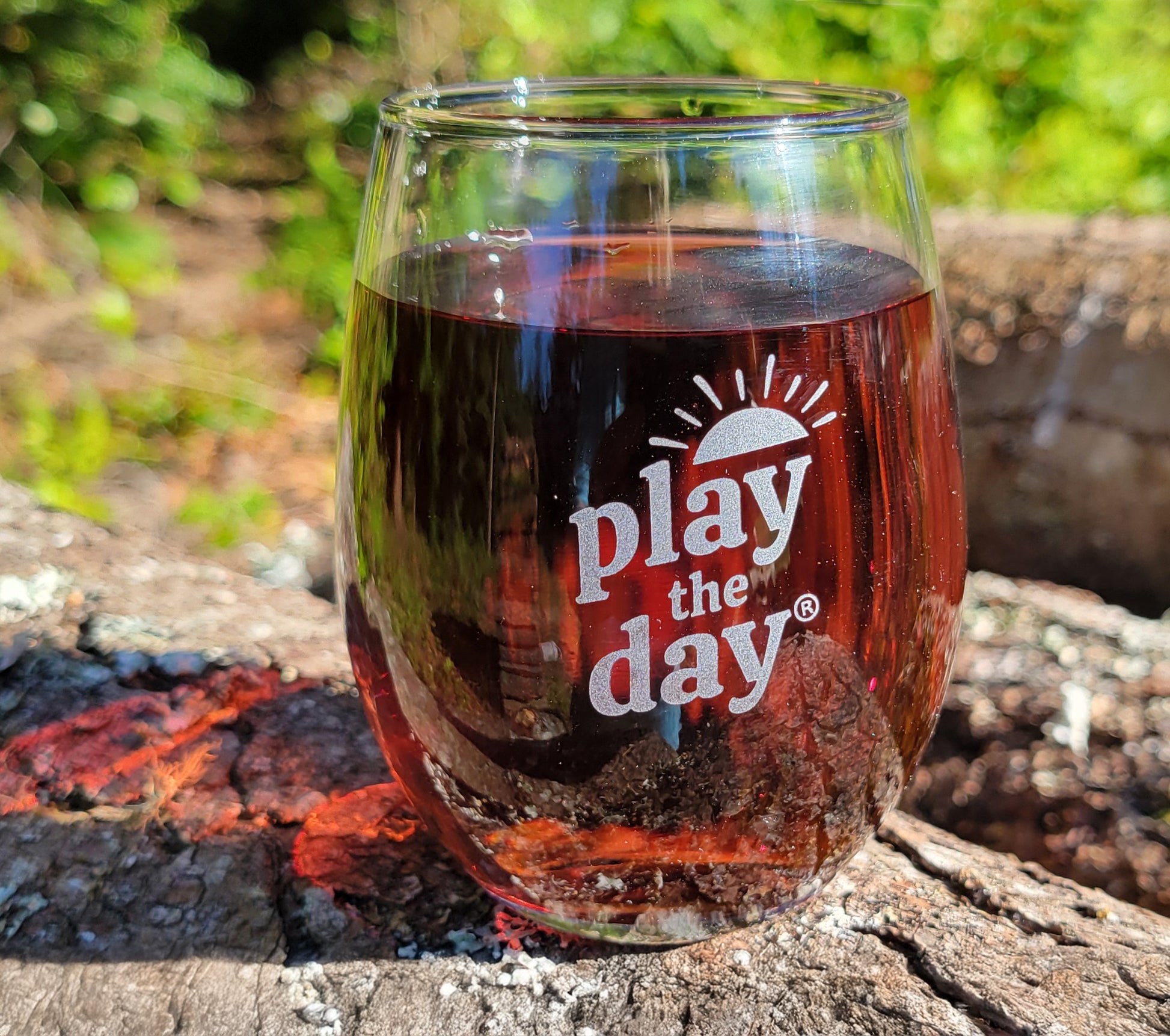 Play the Day® stemless glass filled with a dark beverage, resting on a sunlit log outdoors.