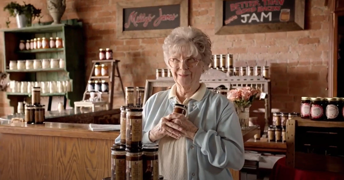 Sydney Dickinson smiling and holding a jar of jam in a rustic store, surrounded by shelves of preserves.